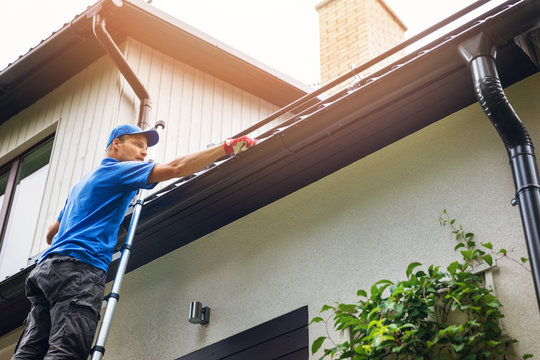 A man in a blue shirt and cap stands on a ladder, cleaning gutters of a house